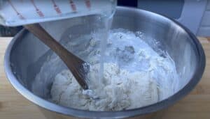 Mixing water into flour in a metal bowl to make bread dough, with a wooden spoon stirring the ingredients.